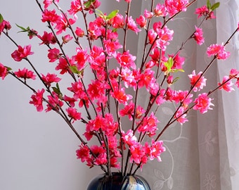 May include: A vibrant floral arrangement featuring bright pink blossoms and green leaves. The flowers are arranged in a blue vase with a glossy finish, creating a striking contrast. The branches are long and slender, adding height and visual interest.