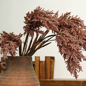 Puede incluir: Un ramo de hierba de la pampa seca de color rosa en una maceta de barro marrón rústica con dos asas. La maceta está sobre una estantería de madera con una pila de bloques de madera detrás.