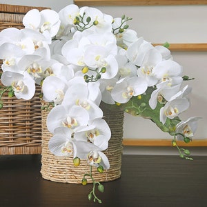以下が含まれることがあります： 籐製の編まれた花瓶に白い蘭の花の飾り付け。花は、花が外側を向くように、滝のように配置されています。花瓶は自然な茶色で、円筒形をしています。