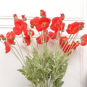 Könnte beinhalten: Ein Strauß künstlicher roter Mohnblumen mit grünen Stielen und Blättern. Die Blumen sind in einer Traube angeordnet, wobei einige Blüten zur Kamera zeigen und andere nach außen zeigen.
