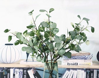 May include: A bouquet of artificial eucalyptus stems with small white flowers. The stems are arranged in a clear glass vase with a green base. The vase is sitting on a shelf with books.