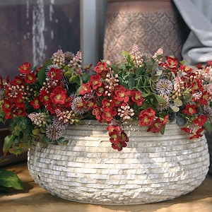 May include: A white ceramic planter with a textured surface, filled with a bouquet of red and pink flowers. The flowers are arranged in a circular shape and are spilling over the edges of the planter.
