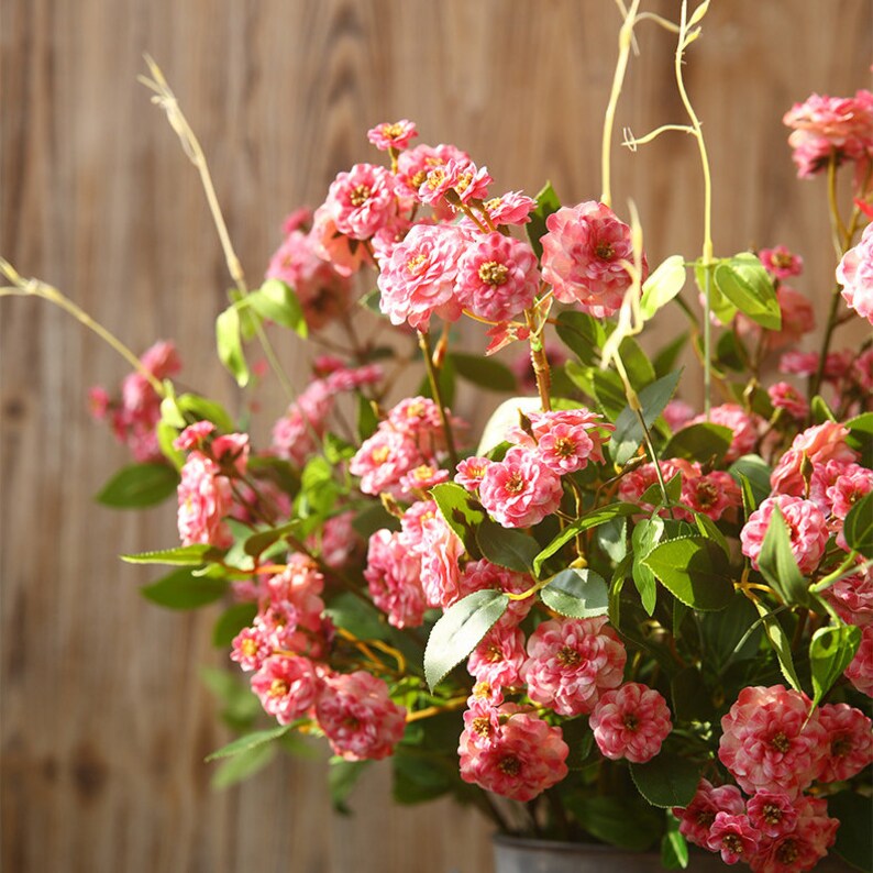 May include: A bouquet of pink artificial flowers with green leaves. The flowers are clustered together and have a delicate, ruffled appearance.
