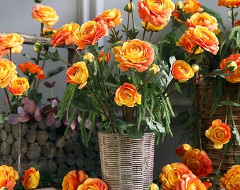 May include: A bouquet of artificial orange and yellow ranunculus flowers in a woven basket vase. The flowers are arranged in a vase with a woven texture.