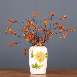 Peut inclure: Un vase en céramique blanche avec un motif de tournesol et un bord doré. Le vase est rempli de branches artificielles orange et jaunes.