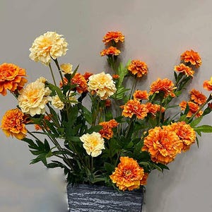 Puede incluir: Un ramo de flores artificiales de cal&eacute;ndula en tonos naranja y amarillo. Las flores est&aacute;n dispuestas en un jarr&oacute;n de piedra gris.