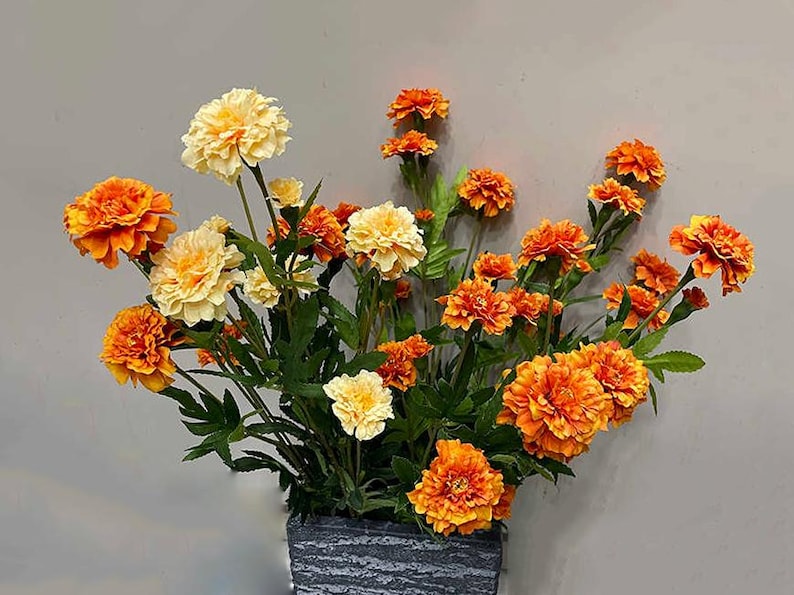 Puede incluir: Un ramo de flores artificiales de cal&eacute;ndula en tonos naranja y amarillo. Las flores est&aacute;n dispuestas en un jarr&oacute;n de piedra gris.