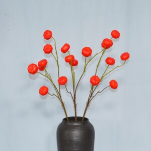 Puede incluir: Un jarrón gris carbón oscuro que contiene un arreglo de ramas decorativas con múltiples bayas pequeñas, redondas y de color naranja brillante con forma de calabaza. Los tallos son delgados y marrones, ramificándose.