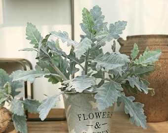 May include: An artificial plant with silvery-green leaves potted in a white metal pot with the text "Flowers & Garden" printed on the front.