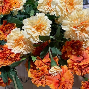 Puede incluir: Un ramo de flores artificiales con flores de color naranja y crema. Las flores tienen una apariencia similar a la de los claveles y est&aacute;n dispuestas en un racimo.