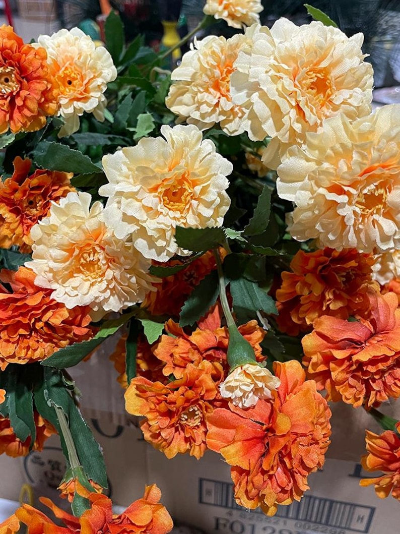 Puede incluir: Un ramo de flores artificiales con flores de color naranja y crema. Las flores tienen una apariencia similar a la de los claveles y est&aacute;n dispuestas en un racimo.