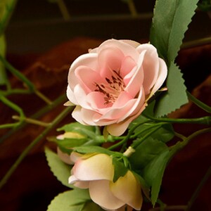 Puede incluir: Primer plano de una flor artificial rosa y blanca con hojas verdes. La flor tiene un aspecto delicado y en capas y es una representaci&oacute;n realista de una rosa real.