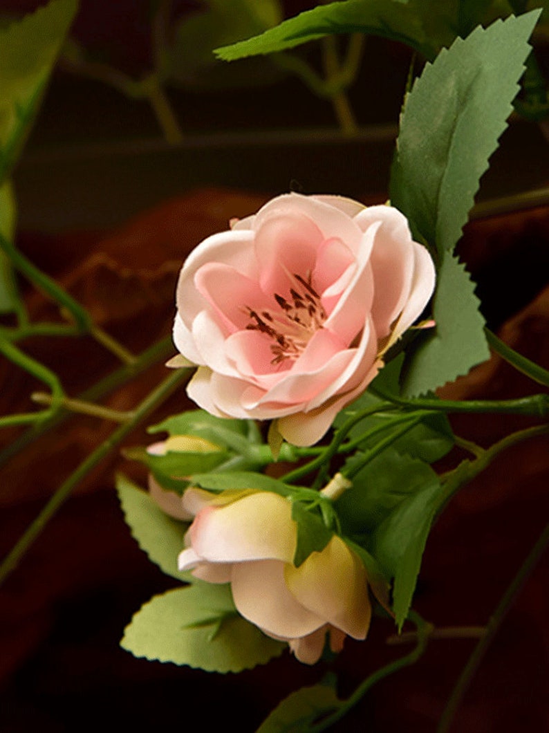 Puede incluir: Primer plano de una flor artificial rosa y blanca con hojas verdes. La flor tiene un aspecto delicado y en capas y es una representaci&oacute;n realista de una rosa real.