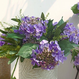 Op de afbeelding: Een boeket paarse hortensia bloemen in een witte vaas met een getextureerd patroon. De bloemen zijn in een tros gerangschikt met groene bladeren.