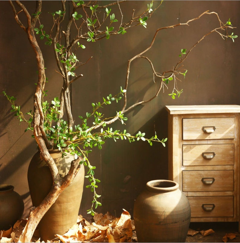 May include: A rustic brown ceramic vase with a dried branch and green leaves. The vase is sitting on a table with a wooden chest of drawers in the background.