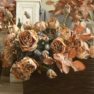 Op de afbeelding: Een boeket gedroogde rozen in tinten bruin en beige, gerangschikt in een geweven rieten mand. De rozen zijn bij elkaar gegroepeerd, wat een rustieke en elegante presentatie creëert.