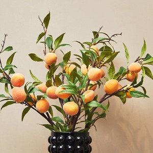 Peut inclure: Un vase noir texturé contenant une branche de fleurs de pêcher artificielles avec des feuilles vertes et des fruits orange.