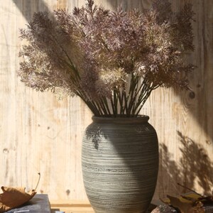May include: A bouquet of dried, light brown and purple pampas grass in a gray, textured ceramic vase. The vase has a wide, round base and a narrow neck.