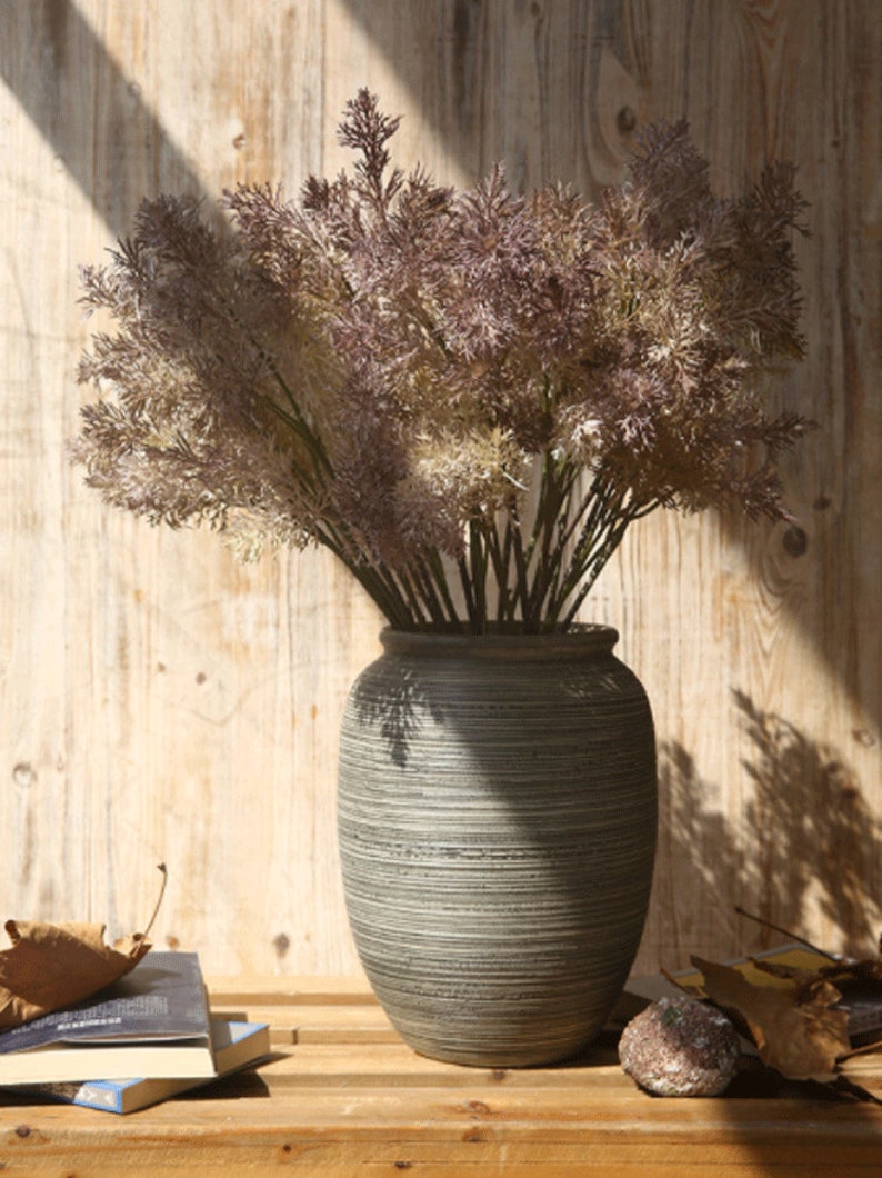 May include: A bouquet of dried, light brown and purple pampas grass in a gray, textured ceramic vase. The vase has a wide, round base and a narrow neck.