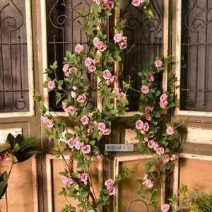 Puede incluir: Una larga vid de rosas artificiales de color rosa con hojas verdes est&aacute; colgada sobre una puerta de madera con una reja de metal negro. La vid cuelga por el lado de la puerta y est&aacute; enfocada. La puerta est&aacute; desenfocada.