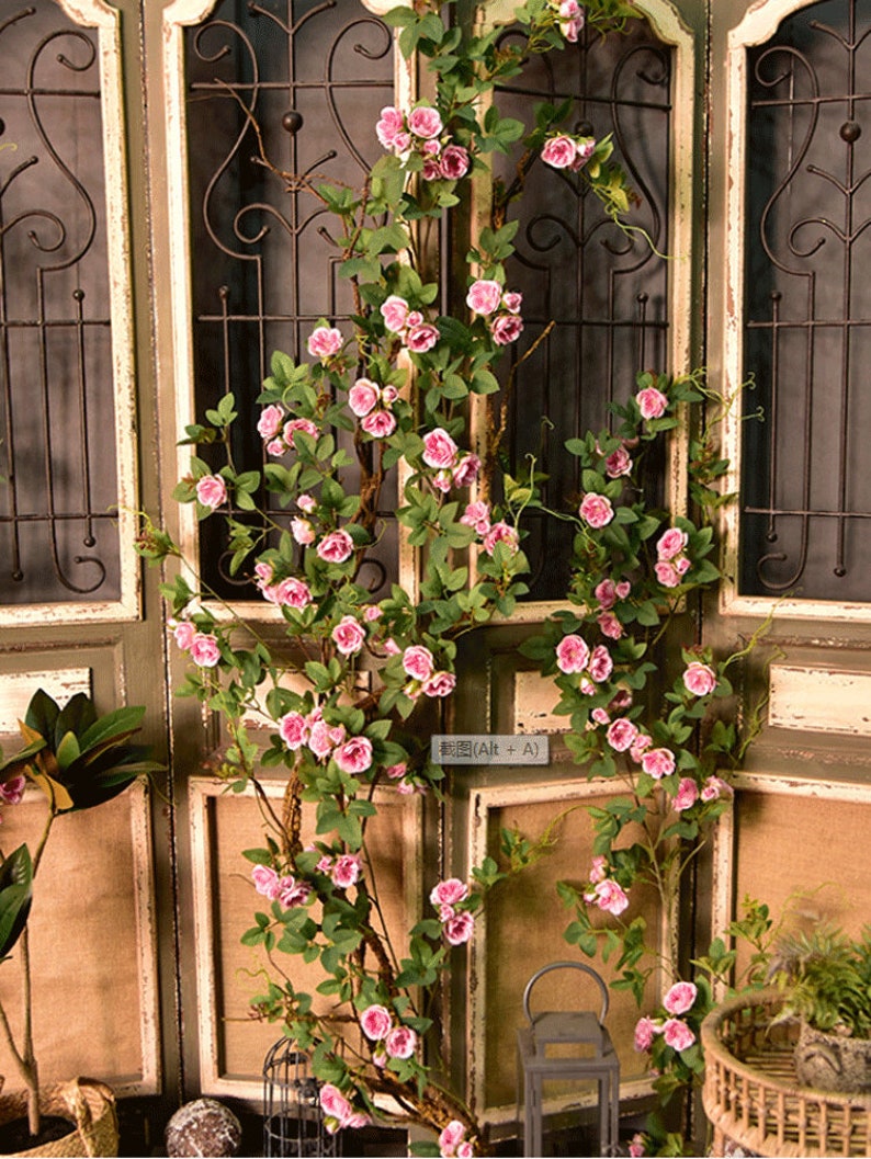 Puede incluir: Una larga vid de rosas artificiales de color rosa con hojas verdes est&aacute; colgada sobre una puerta de madera con una reja de metal negro. La vid cuelga por el lado de la puerta y est&aacute; enfocada. La puerta est&aacute; desenfocada.