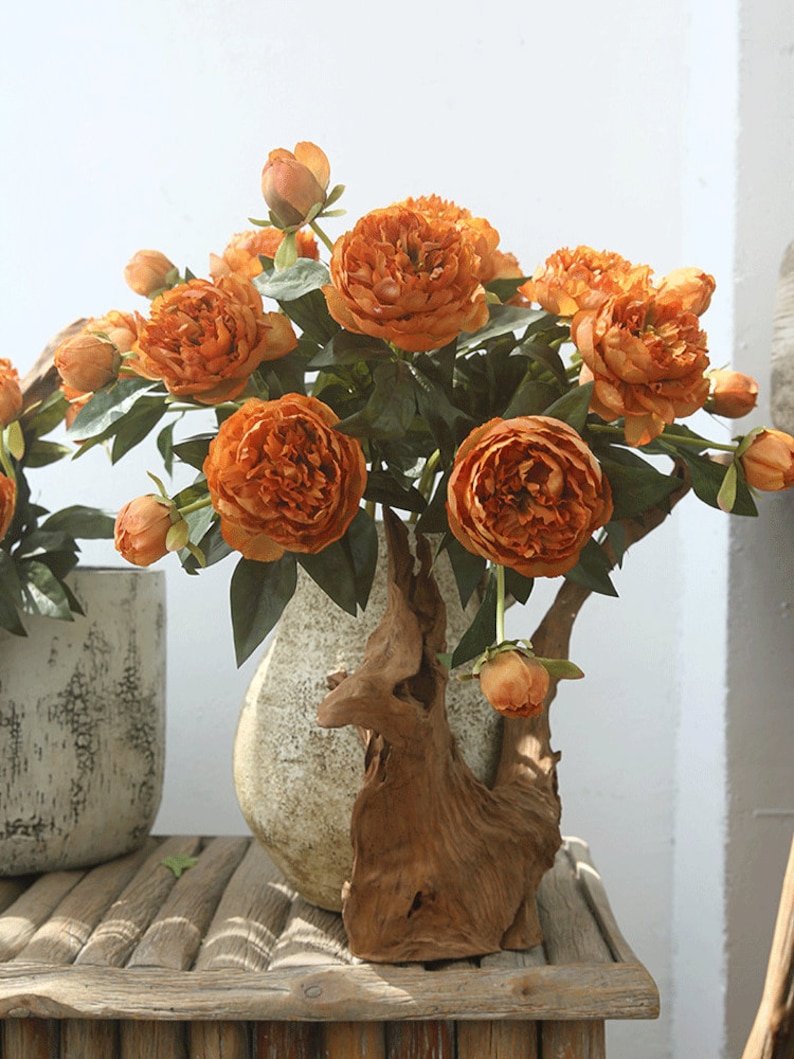 May include: A bouquet of artificial orange peonies in a rustic wooden vase. The flowers are arranged in a natural, flowing style, with some buds still closed. The vase is made of driftwood and has a unique, organic shape.