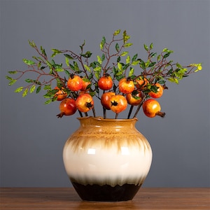 Może przedstawiać: Ceramiczny wazon z brązową, białą i czarną glazurą wypełniony jest bukietem sztucznych gałązek granatu. Gałązki mają zielone liście i czerwone oraz pomarańczowe granaty.