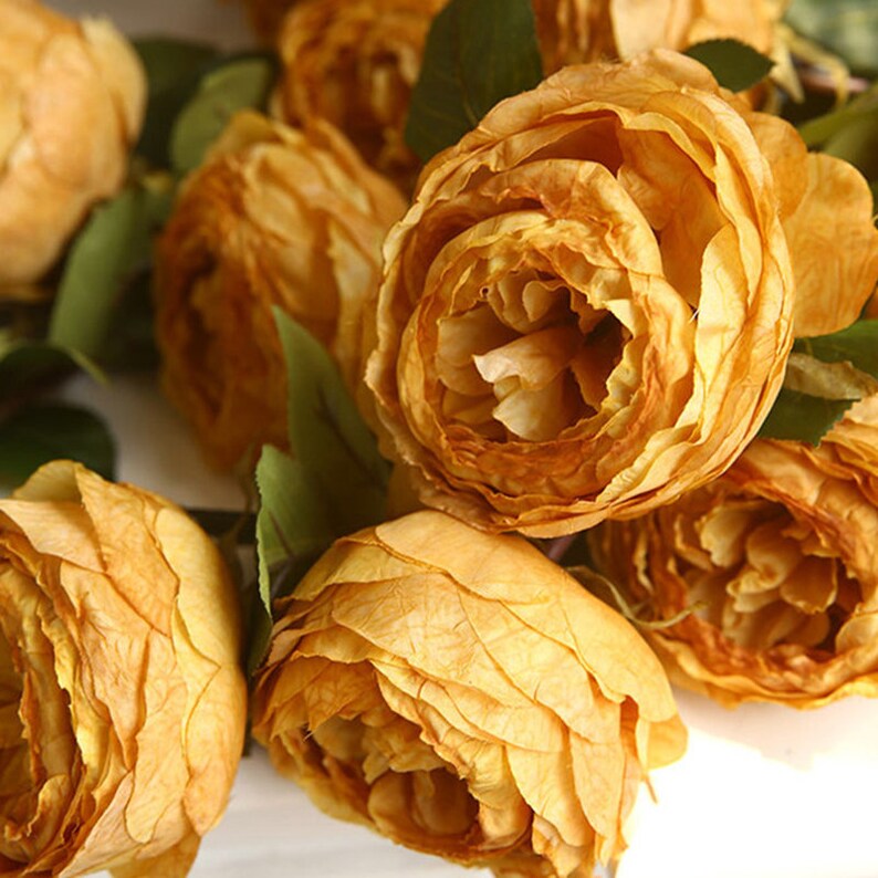 Puede incluir: Un ramo de rosas artificiales de color amarillo intenso y apagado. Las rosas est&aacute;n dispuestas en un racimo, con los p&eacute;talos superpuestos y creando un aspecto suave y texturizado.