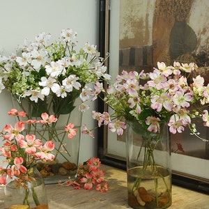 Puede incluir: Tres jarrones de vidrio llenos de flores artificiales. Los jarrones son de vidrio transparente y las flores son blancas, rosas y naranjas.