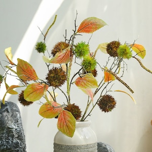May include: A bouquet of faux autumn leaves and burrs in shades of green, brown, and orange arranged in a white vase.
