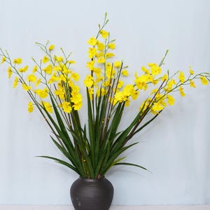 Puede incluir: Arreglo floral decorativo con flores amarillas brillantes, tallos largos y delgados y hojas verdes. Las flores están dispuestas en un jarrón gris oscuro, creando un contraste llamativo. El jarrón mide unos 30 cm de alto.
