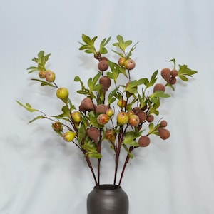 May include: A decorative artificial branch arrangement featuring numerous small, round fruits in shades of brown, yellow, and green, interspersed with vibrant green leaves on thin, dark brown stems, all contained within a dark grey cylindrical vase.