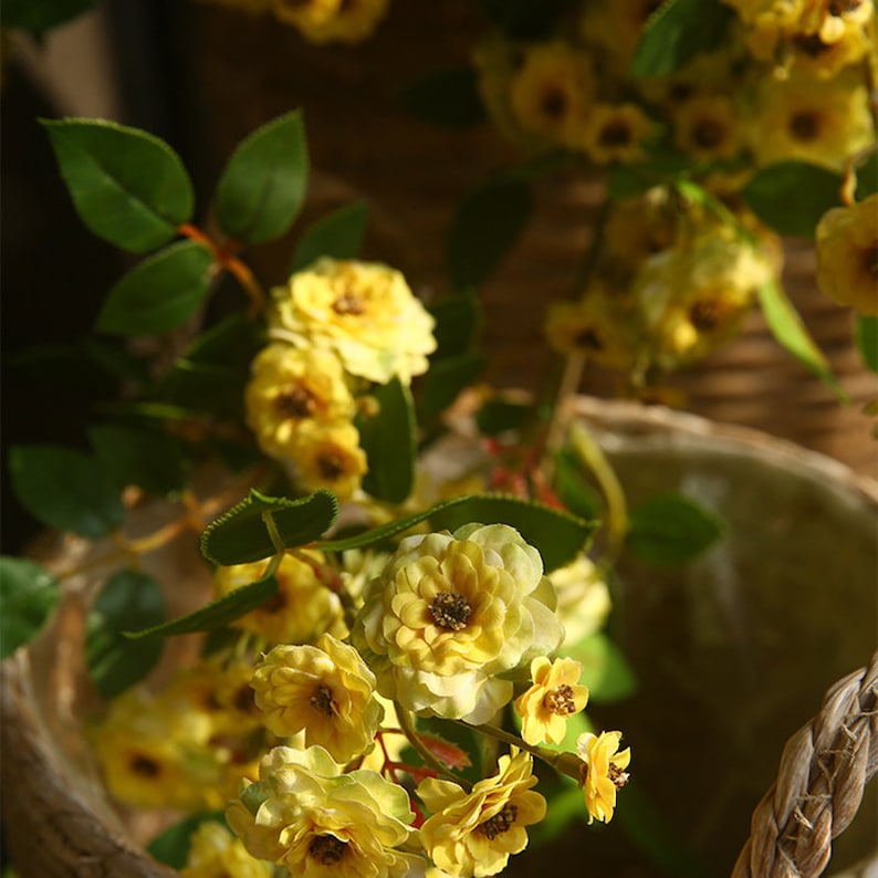 May include: A bouquet of yellow artificial flowers with green leaves in a brown wicker basket. The flowers are arranged in a cascading style, with the blooms at the top and the leaves trailing down the sides.