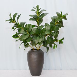 May include: A dark brown, textured vase holds a lush arrangement of artificial greenery. The leaves are a vibrant green, with a mix of large, rounded leaves and smaller, pointed ones. The vase has a simple, elegant shape.