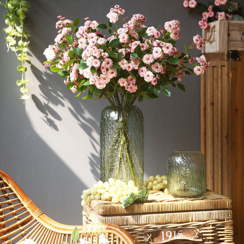 May include: A bouquet of pink flowers in a clear textured glass vase. The vase is sitting on a wooden table with a wicker basket and a small glass vase.