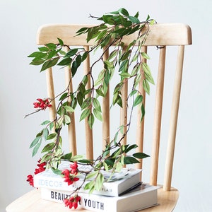 May include: A light wooden chair with a green leafy garland and red berries draped over it. The garland rests on a stack of books with the words "DECOR" and "BEAUTY" visible. The chair has a simple, classic design.