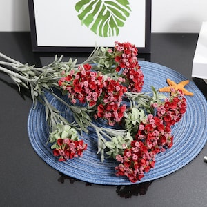 May include: A bouquet of artificial red flowers with green leaves. The flowers are arranged in a cluster and have a delicate, realistic appearance. The bouquet is displayed on a blue and white woven placemat.