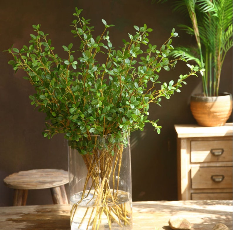 May include: A vase filled with artificial green leafy branches. The vase is clear glass and the branches are arranged in a bouquet.