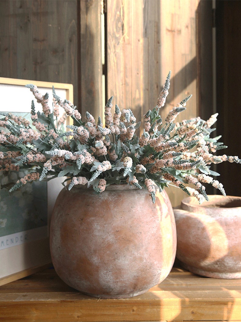 Puede incluir: Un ramo de flores artificiales de color rosa p&aacute;lido y verde en una maceta de terracota. La maceta tiene un acabado blanco desgastado.