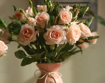 May include: A bouquet of peach-colored artificial roses in a pink vase with a floral pattern. The vase has a string tied around the neck.
