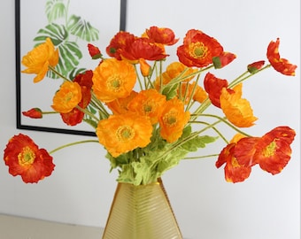 Könnte beinhalten: Ein Strauß künstlicher Mohnblumen in Orange- und Rottönen. Die Blumen sind in einer gelben Vase mit strukturierter Oberfläche angeordnet.