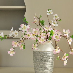 May include: A white vase with a gray textured pattern holds a bouquet of pink and white artificial flowers. The flowers have delicate petals and are arranged in a natural, flowing style.
