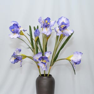 May include: A bouquet of artificial irises in a dark brown vase. The flowers have purple and white petals with yellow centers and green stems. The arrangement is set against a plain white background.
