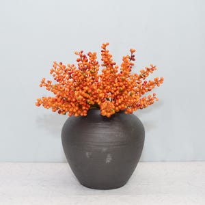 Może przedstawiać: Ciemnoszary, teksturowany ceramiczny wazon zawiera żywą kompozycję pomarańczowych jagód. Wazon ma zaokrąglony kształt i stoi na białej powierzchni, tworząc uderzający kontrast z jasnymi jagodami.
