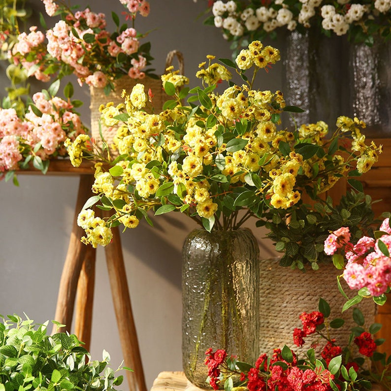 May include: A bouquet of yellow artificial flowers in a clear glass vase with a textured surface. The vase is on a wooden table with a brown wooden stool behind it. There are other artificial flowers in the background.
