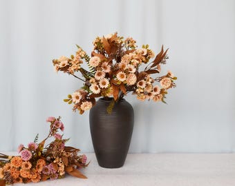 Könnte beinhalten: Eine dunkelbraune, strukturierte Vase enthält ein großes Arrangement künstlicher Blumen. Die Blumen sind in Pfirsich-, Creme- und Brauntönen gehalten, mit grünen Akzenten. Ein kleinerer Strauß liegt auf der Oberfläche.
