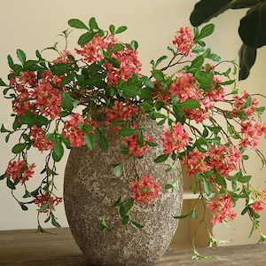 Könnte beinhalten: Ein Strauß künstlicher rosa Blumen mit grünen Blättern in einer strukturierten grauen Keramikvase.
