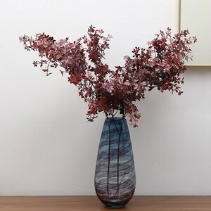 以下が含まれることがあります： 波打つ模様の入った青と白のガラスの花瓶に、乾燥した赤い葉のブーケが活けられています。