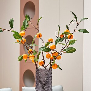 May include: A vase with a textured gray surface holds a branch with green leaves and small orange fruit.