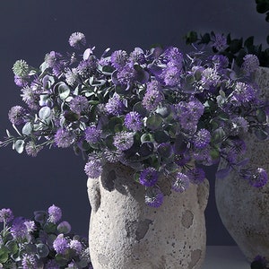 Peut inclure: Un bouquet de fleurs de trèfle violet dans un vase en pierre rustique gris. Les fleurs sont disposées dans un style lâche et naturel, avec quelques tiges qui débordent du vase.